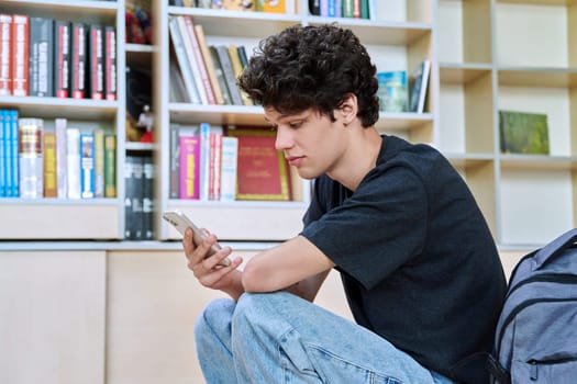 Relaxed handsome young male college student using smartphone, sitting inside library classroom. Teenager guy 19-20 years old, education, internet technology, mobile applications, lifestyle, youth