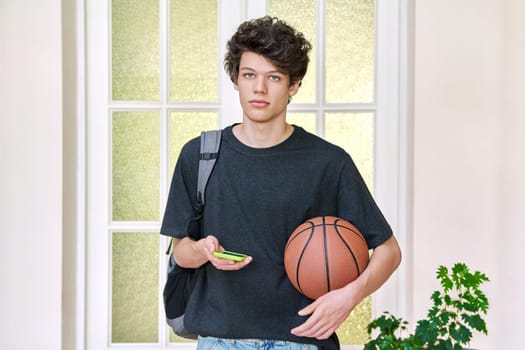 Portrait of young male with backpack, basketball ball and smartphone. Handsome curly 19, 20 year old teen student looking at camera inside college. Lifestyle, youth concept