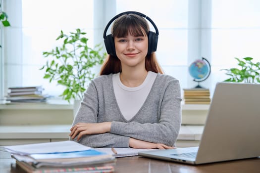 Portrait of teenage college student wearing headphones, sitting at desk, smiling and looking at camera. Education, training, youth 18,19, 20 years old