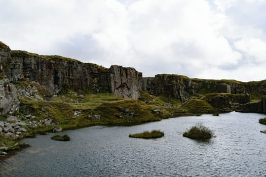 A serene landscape featuring a rocky cliffside with patches of green vegetation, overlooking a calm body of water. The sky is partly cloudy, adding a dramatic effect to the scene.