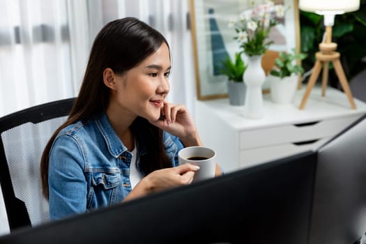 Working young beautiful asian creative manager with glasses focusing on pc while drinking hot coffee, creating with website design on social media with blue jeans shirt at home office. Stratagem.