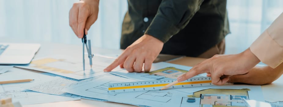 A cropped image of professional engineer using divider to measure blueprint at meeting table with blueprint, laptop and architectural equipment scatter around. Closeup. Delineation.