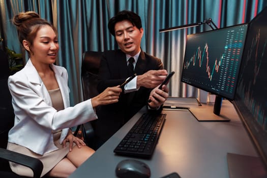 Asian businessman showing stock market application on mobile phone to partner pointing by pen, comparing financial digital investment database on computer screen at neon light at night time. Infobahn.