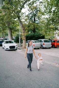 Smiling mother and her little girl walk holding hands along the road past parked cars. High quality photo