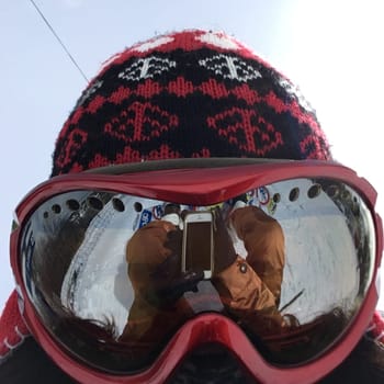 Close-up of a person wearing ski goggles and a red and black knit hat, with a reflection of a snowy landscape and a phone in the goggles.