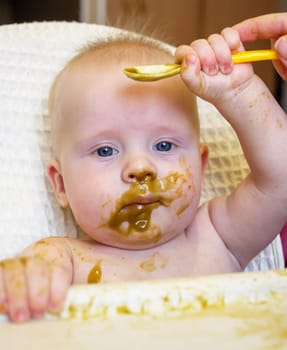 A cute baby with blue eyes has broccoli puree smeared all over their face. It looks like the baby is enjoying their meal while sitting at home.