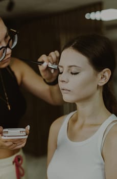 One young handsome Caucasian makeup artist applies foundation powder with a fluffy brush to the temples of a girl sitting in a chair early in the morning in a beauty salon, close-up side view. Step by step.