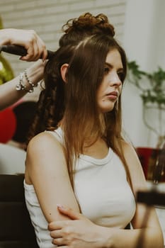 Portrait of a young beautiful Caucasian brunette girl sitting in a hairdresser's shop, where the master is curling her long hair on a flat iron, close-up view of the side.