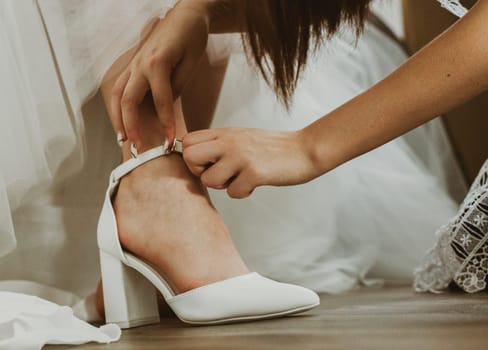 The hand of an unrecognizable bridesmaid helps to put white wedding shoes on the bride's leg, close-up side view.
