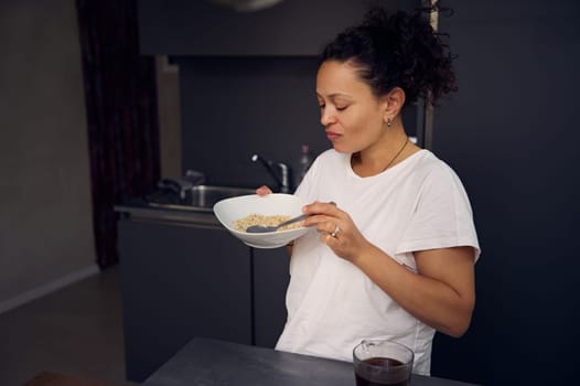 Happy African American woman in pajamas, holding a muesli bowl in the home kitchen. Healthy lifestyle. People. Diet. Food consumerism.