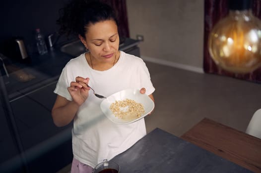 View from above of a happy Latina African American woman in pajamas, holding a muesli bowl in the home kitchen. Healthy lifestyle. People. Diet. Food consumerism.