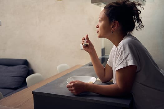 Side portrait of a curly haired multi ethnic woman eating oat flakes for breakfast, standing at kitchen island, dressed in white t-shirt. People. Food and drink consumerism. Healthy diet and slimming