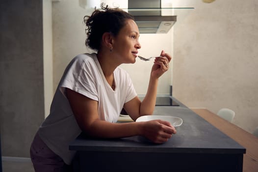 Authentic side portrait of a curly haired mixed race woman eating oat flakes for breakfast, standing at kitchen island, dressed in white t-shirt. People. Food and drink consumerism