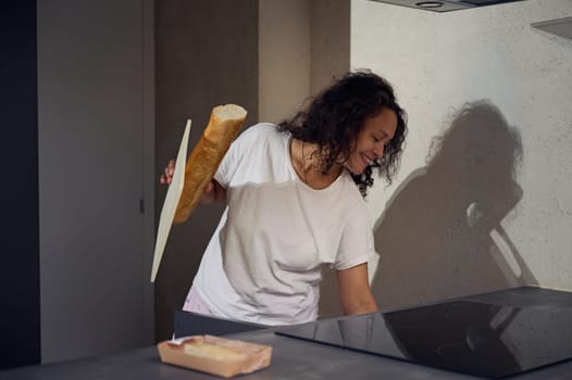 Multi ethnic young adult woman smiling broadly, holding a loaf of bread, standing at kitchen counter at home, preparing breakfast in the morning. People. Morning routine. Domestic life