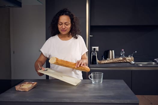 Authentic portrait of a young woman holding a loaf of bread and cutting board, standing at kitchen counter at home, starting prepare breakfast in the morning. Domestic life. People and lifestyle