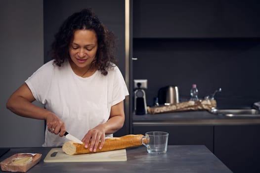 Pretty woman in white t-shirt, standing at kitchen counter, holding a kitchen knife nd cutting bread on the cutting board, preparing breakfast in the home kitchen in the morning