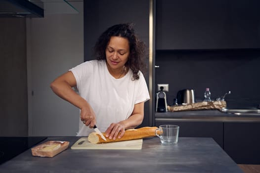 Mixed race beautiful woman in white t-shirt, standing at kitchen counter, holding knife and slicing a loaf of French baguette on a cutting board, preparing breakfast in the home kitchen in the morning