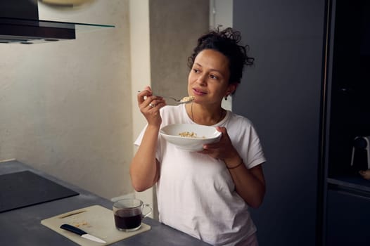 Young pretty woman in white t-shirt, eating a bowl of muesli at home. People. Food consumerism. Diet and slimming concept. Healthy lifestyle