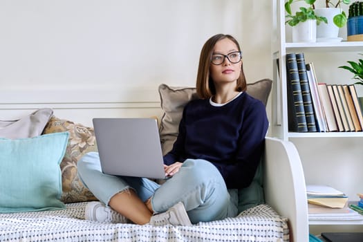 Young woman in glasses working at home, sitting on couch using laptop computer. Freelancing, training, education, remote work, leisure, internet online technology applications apps concept