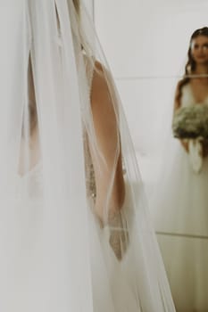 Portrait of one beautiful young Caucasian recognizable brunette bride holding a bouquet of white boutonnieres and looking in the mirror on the wardrobe in the bedroom, side view close-up with selective focus.