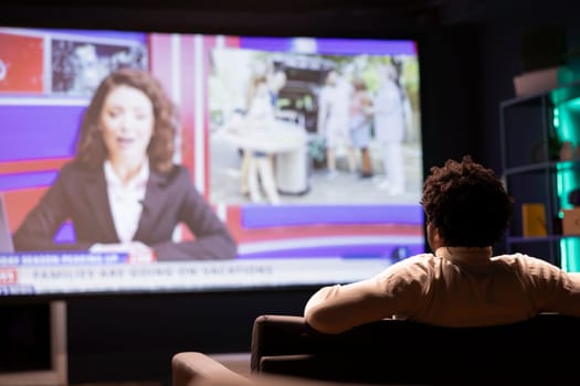 Man sitting on couch in front of ultrawide smart TV displaying news program broadcasting. Person watching big television screen showing VOD channel with newscast coverage