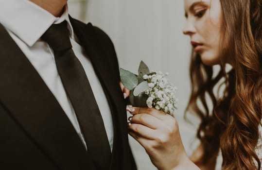 Portrait of one young beautiful stylish Caucasian brunette bride inserting a mini bouquet of boutonnieres into the groom's chest pocket in a dark suit, close-up side view. Wedding traditions concept.