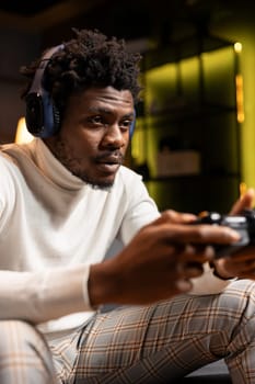 African american man playing videogame in yellow neon lit apartment, holding controller. Gamer participating in PvP online multiplayer game using console system and headphones for immersive experience