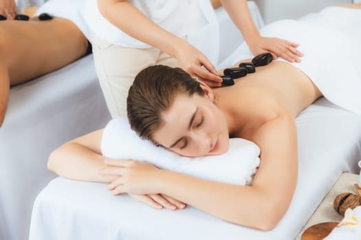 Hot stone massage at spa salon in luxury resort with day light serenity ambient, blissful couple customer enjoying spa basalt stone massage glide over body with soothing warmth. Quiescent