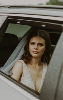 Portrait of one young beautiful stylish Caucasian brunette bride with a serious emotion on her face sits inside a car and looks at the camera through an open window on a rainy cloudy day, close-up side view.