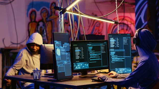 Hackers in secret bunker with graffiti walls writing code that spoofs their location, tricking cops. Rogue programmers running script pinging wrong location to cybercriminal law enforcement