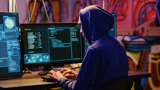 Hacker in graffiti painted hideaway base writing lines of code on computer, developing malware that get past security systems. Rogue programmer uses PC in bunker, running hacking script