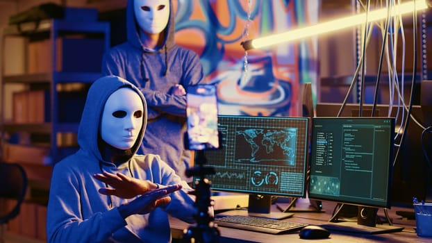 Masked hackers filming ransom video in secret HQ, threatening to release stolen data publicly if payout demands are not met. Cybercriminals using smartphone to record blackmail footage