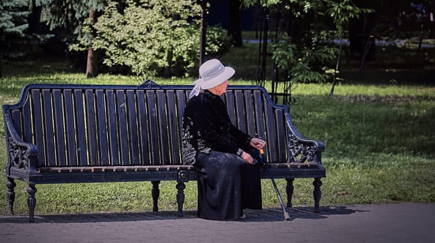 An elderly woman wearing a black dress and white hat sits on a park bench, surrounded by lush trees. The serene expression on her face suggests that she is enjoying the peace and quiet of the park.