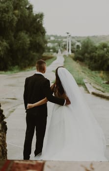 Portrait of one young beautiful stylish Caucasian bride and groom standing from behind and hugging each other, looking into the distance at the road leading forward on a rainy cloudy day, side view close-up with depth of field.