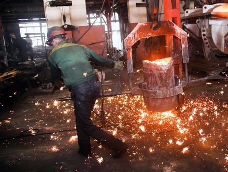 Steel mill worker wearing safety gear and a hard hat carefully pours glowing molten metal into a mold, creating sparks as industrial machinery hums around them in a busy factory setting.