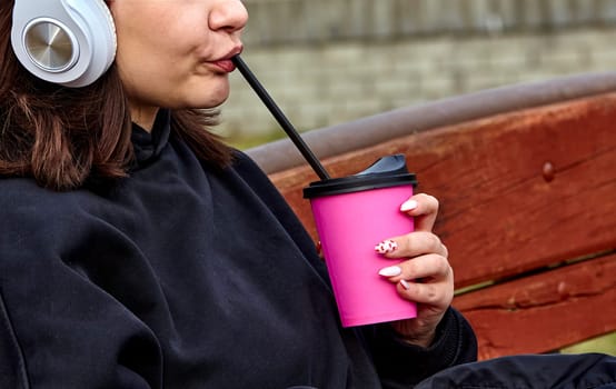 A young woman with long brown hair and white headphones is drinking from a pink cup with a straw. She is wearing a black sweatshirt and has her nails painted pink with white polka dots.