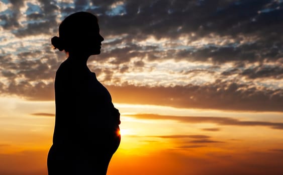 The silhouette of a pregnant woman at sunset with her hands on her belly. The sky is ablaze with color, and the clouds are forming a beautiful pattern. A truly stunning and peaceful scene.