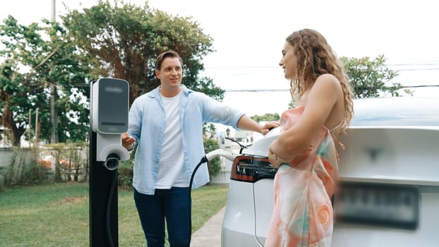 Happy and lovely couple with eco-friendly conscious recharging electric vehicle from EV charging station. EV car technology utilized as alternative transportation for future sustainability. Synchronos