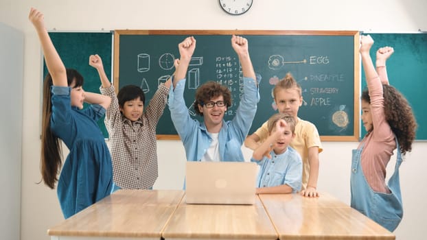 Teacher and multicultural student looking at laptop while celebrate success project and put hands in the air. Group of children smiling to camera while standing at blackboard in classroom. Pedagogy.