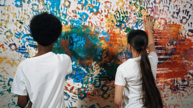 Back view of young diverse children paint colorful stained wall with hand. Attractive highschool student paint the wall with hand print while holding color palette. Creative activity. Edification.