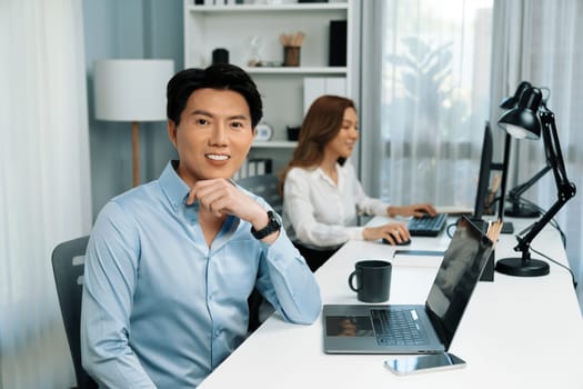 Profile smiling of smart Asian businessman looking camera to pose at modern office. Blurry background woman colleague working on pc to check email marketing plan analysis report at desk. Infobahn.