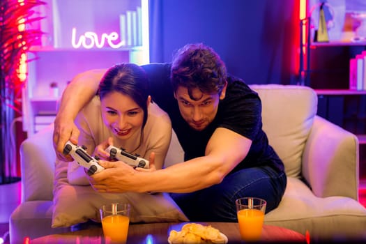 Couple joyful funny players playing video game on TV using joysticks, fighting winner trying to disturb when focusing level challenge in red neon light studio at comfy living home place. Postulate.