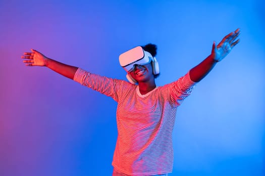 Joyful African woman with VR headset raised hands in excitement on neon studio lighting in virtual reality entertainment with metaverse new world 3D hologram technology flower air fresh. Contrivance.