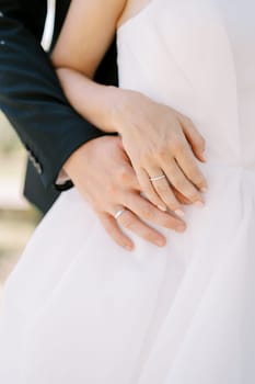 Groom hugs bride from behind, placing her hand on his hand. Cropped. Faceless. High quality photo