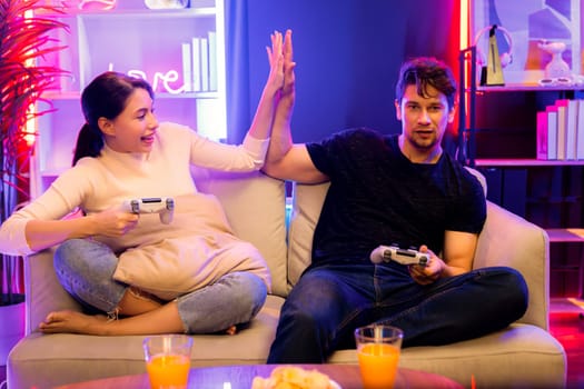 Couple joyful of player video game on TV using joysticks making high five of winner together on competition gaming in studio room in red blue neon light bulb at comfy living home place. Postulate.