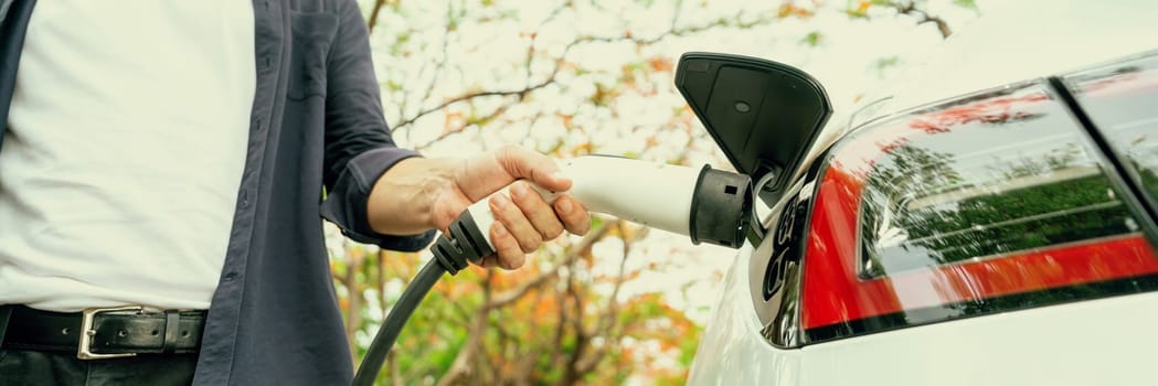 Panorama banner man recharging battery for electric car during autumnal road trip travel EV car in autumnal forest. Eco friendly travel on vacation during autumn with electric vehicle. Exalt