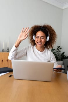 Vertical portrait of young multiracial hispanic woman waving hand on a video call at home office using wireless headphones and laptop. Working at home concept.
