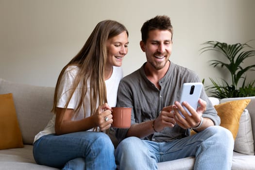 Smiling young caucasian couple relaxing at home using phone together looking social media. Lifestyle concept.