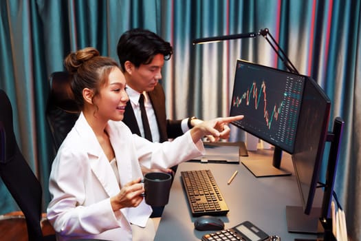 Smiling businesswoman in white suit pointing on dynamic stock exchange statistic on pc screens holding coffee cup, comparing data sitting by coworker searching folder at night neon office. Infobahn.