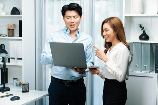 Smiling Asian coworkers standing while talking planning marketing report project on laptop casual day at modern office. Analyzing successful customer need in new social media online channel. Infobahn.